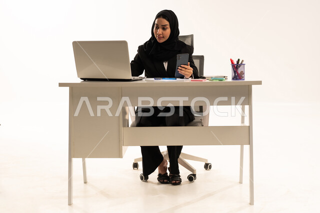 إرسال بريد الكتروني عبر الحاسوب, التواصل مع العملاء عن بعد, استخدام التقنيات الحديثة و المتطورة في العمل, بورتريه لامرأة عربية سعودية خليجية محجبة ترتدي البدلة الرسمية السوداء تستخدم اللابتوب و تحمل الموبايل, خلفية بيضاء