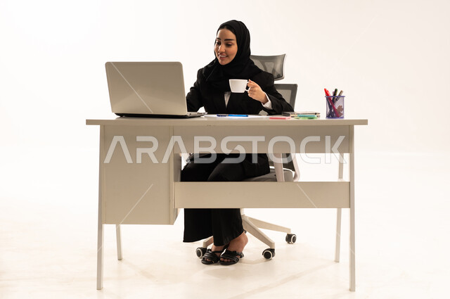 احتساء المشروب المفضل في المكتب، ادارة الأعمال وانجاز المهام المطلوبة، العمل عن بعد، مهنة ووظيفة نسائية، بورتريه لامرأة عربية سعودية خليجية محجبة ترتدي البدلة الرسمية السوداء تستخدم اللابتوب و الموبايل و تشرب مشروب دافئ, خلفية بيضاء