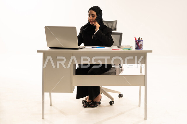 التركيز والتفاني والعمل الجاد، استخدام التقنيات الحديثة والتكنولوجية في الأعمال الإدارية والمكتبية، بورتريه لامرأة عربية سعودية خليجية محجبة تجلس خلف المكتب ترتدي البدلة الرسمية السوداء و تستخدم اللابتوب أثناء إجراء مكالمة هاتفية عبر الموبايل, خلفية بيضاء