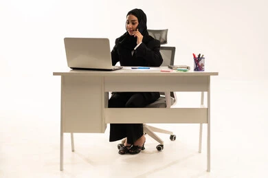 Focus, dedication, and hard work; the use of modern techniques and technology in administrative and office work; a portrait of a veiled Saudi Arabian woman sitting behind a desk wearing a black formal suit and using a laptop while making a phone call on her mobile phone; white background.