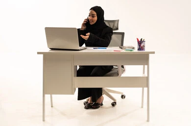 الدرشة والتواصل مع الاصدقاء في وقت العمل، مهن ووظائف مكتبية سعودية،  بورتريه لامرأة عربية سعودية خليجية محجبة تجلس خلف المكتب ترتدي البدلة الرسمية السوداء و تستخدم اللابتوب أثناء إجراء مكالمة هاتفية عبر الموبايل, خلفية بيضاء