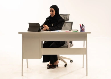 Focus on completing tasks, integrating modern technologies into the administrative and office field; a portrait of a veiled Saudi Arabian Gulf woman sitting behind a desk wearing a black formal suit and using a tablet, white background.