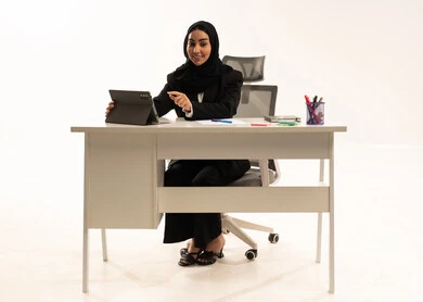شركات ومكاتب خليجية في السعودية, جلسه عمل عن بعد، التركيز في إنجاز المهام، دمج التقنيات الحديثة بالمجال الإداري والمكتبي، بورتريه لامرأة عربية سعودية خليجية محجبة مبتسمة تجلس خلف المكتب ترتدي البدلة الرسمية السوداء و تستخدم التابلت, خلفية بيضاء