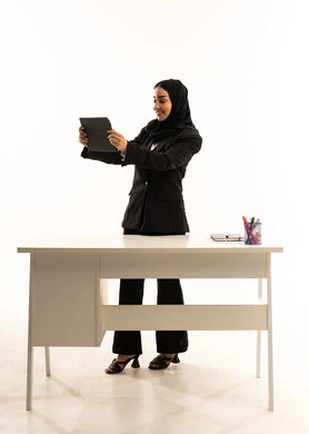 المنهاج المتطور والتعليم الالكتروني, جودة أساليب التعلم الفعال، بورتريه لمعلمه عربيه سعوديه خليجيه مبتسمة محجبة ترتدي البدله الرسميه السوداء تقف امام مكتب ابيض و تحمل جهاز التابلت, خلفية بيضاء