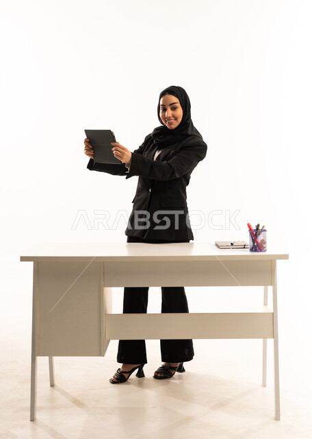 The quality of effective learning methods, the advanced curriculum, and e-learning; a portrait of a smiling, veiled Saudi Arabian teacher wearing a black formal suit, standing in front of a white desk and holding a tablet, white background.