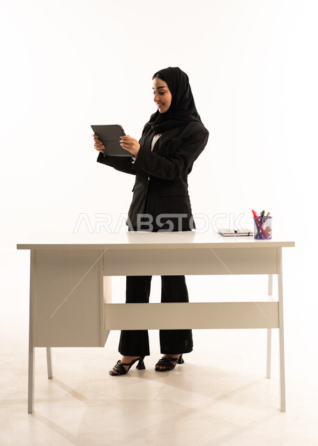 التعلم عن بعد بواسطة الانترنت، مفهوم الشرح والتدريس, اهمية التعليم في المملكة العربية السعودية، بورتريه لمعلمه عربيه سعوديه خليجيه مبتسمة محجبة ترتدي البدله الرسميه السوداء تقف امام مكتب ابيض و تحمل جهاز التابلت, خلفية بيضاء