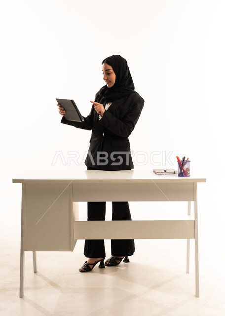 الاهتمام بالتعليم والتطوير, اهمية التعلم في المملكة العربية السعودية، بورتريه لمعلمه عربيه سعوديه خليجيه مبتسمة محجبة ترتدي البدله الرسميه السوداء تقف امام مكتب ابيض و تحمل جهاز التابلت, خلفية بيضاء