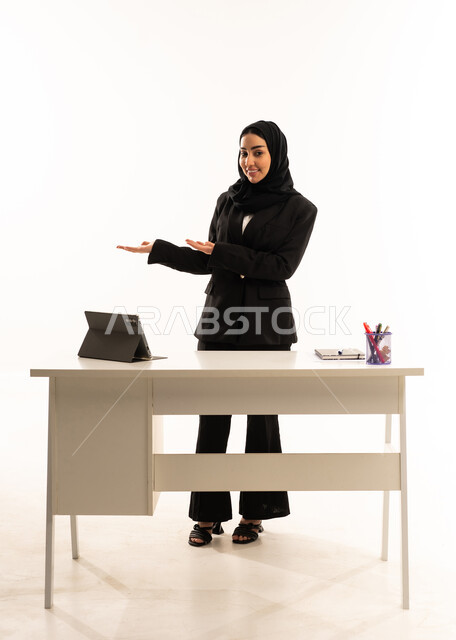 متابعة الدروس التعليمية، الإشارة إلى عنصر ما, بورتريه لمعلمه عربيه سعوديه خليجيه محجبة ترتدي البدله الرسميه السوداء تقف امام مكتب ابيض و تستخدم جهاز التابلت و تشير بيديها على الجانب، شرح موقع شيء معين, خلفية بيضاء