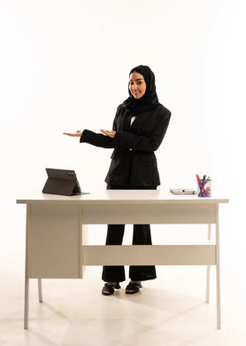 متابعة الدروس التعليمية، الإشارة إلى عنصر ما, بورتريه لمعلمه عربيه سعوديه خليجيه محجبة ترتدي البدله الرسميه السوداء تقف امام مكتب ابيض و تستخدم جهاز التابلت و تشير بيديها على الجانب، شرح موقع شيء معين, خلفية بيضاء