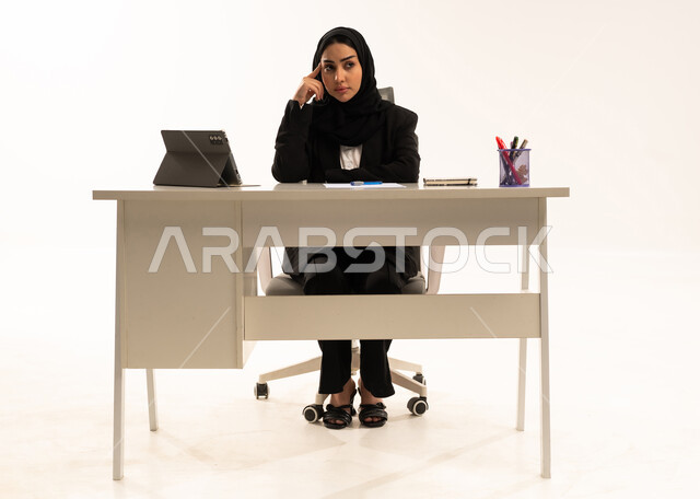 متابعه الأعمال الادارية, بيئة عمل مريحة في الشركات السعودية،  التفكير في الاستراتيجيات والأهداف, بورتريه لامرأة عربية سعودية خليجية محجبة تجلس خلف المكتب ترتدي البدلة الرسمية السوداء و تستخدم التابلت, ايماءات الحيرة و الاندماج, خلفية بيضاء