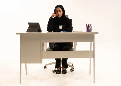 Following up on administrative tasks, a comfortable work environment in Saudi companies, thinking about strategies and goals, a portrait of a veiled Saudi Arabian woman from the Gulf sitting behind a desk wearing a black formal suit and using a tablet, gestures of confusion and absorption, white background