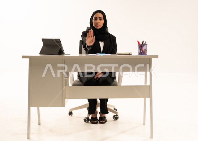 الشعور بالإنزعاج والغضب، الأمر بالتوقف عن فعل شيء ما، بورتريه لامرأة عربية سعودية خليجية محجبة تجلس خلف المكتب ترتدي البدلة الرسمية السوداء و تستخدم التابلت و ترفع يدها بإيماءات تدل على النهي، العمل ضمن مقر الشركة، خلفية بيضاء