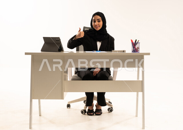 Gestures of approval and acceptance, integrating work with modern technology, a portrait of a veiled Saudi Arabian woman from the Gulf sitting behind a desk wearing a black formal suit, using a tablet and raising her thumbs in a like sign, white background.