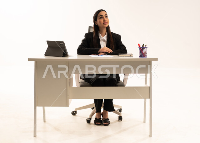 التفكير في مهام العمل, مهن ووظائف مكتبية, دمج التقنيات الحديثة بالمجال الإداري والمكتبي، بورتريه لامرأة عربية سعودية خليجية تجلس خلف المكتب ترتدي البدلة الرسمية السوداء و تستخدم التابلت, خلفية بيضاء