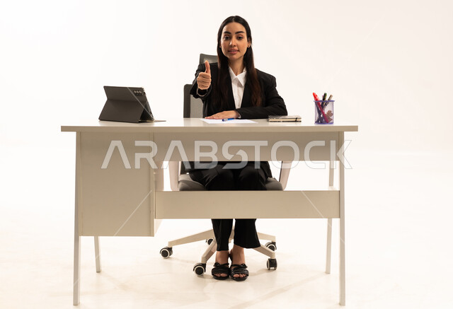الإمتياز والتميز والنجاح، دمج العمل مع التكنولوجيا الحديثة، بورتريه لامرأة عربية سعودية خليجية تجلس خلف المكتب ترتدي البدلة الرسمية السوداء و تستخدم التابلت و ترفع أصابع الإبهام لأعلى بعلامة اللايك, خلفية بيضاء