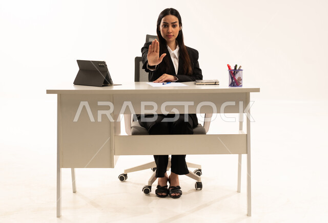 الأمر بالتوقف عن فعل شيء ما، الشعور بالإنزعاج والغضب، بورتريه لامرأة عربية سعودية خليجية تجلس خلف المكتب ترتدي البدلة الرسمية السوداء و تستخدم التابلت و ترفع يدها بإيماءات تدل على النهي، العمل ضمن مقر الشركة، خلفية بيضاء