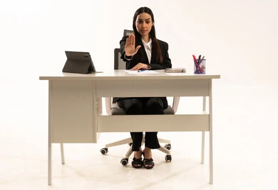 The order to stop doing something, the feeling of annoyance and anger, a portrait of a Saudi Arabian Gulf woman sitting behind a desk wearing a black formal suit, using a tablet and raising her hand in a gesture of prohibition, working within the company premises, white background