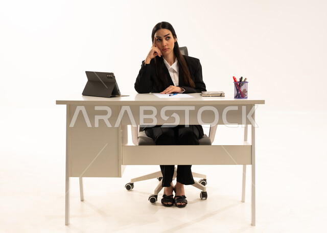العمل ضمن مقر الشركة، التفكير في الاستراتيجيات والأهداف, بورتريه لامرأة عربية سعودية خليجية تجلس خلف المكتب ترتدي البدلة الرسمية السوداء و تستخدم التابلت, ايماءات الحيرة و الاندماج, خلفية بيضاء