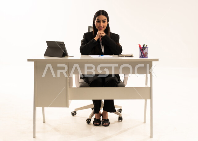 الجلوس في المكتب والنظر إلى الكاميرا, انجاز مهام العمل عبر الانترنت,  بورتريه لامرأة عربية سعودية خليجية تعمل في الشركة ترتدي البدلة الرسمية السوداء و تستخدم التابلت, استخدام التقنيات الحديثة المتطورة، خلفية بيضاء