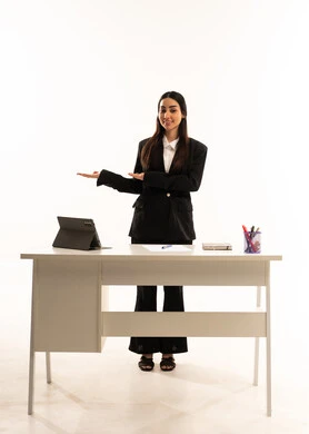 التوضيح والدلالة لمكان محدد, الإشارة إلى عنصر ما,  بورتريه لمعلمه عربيه سعوديه خليجيه ترتدي البدله الرسميه السوداء تقف امام مكتب ابيض و تستخدم جهاز التابلت و تشير بيديها على الجانب، شرح موقع شيء معين, خلفية بيضاء