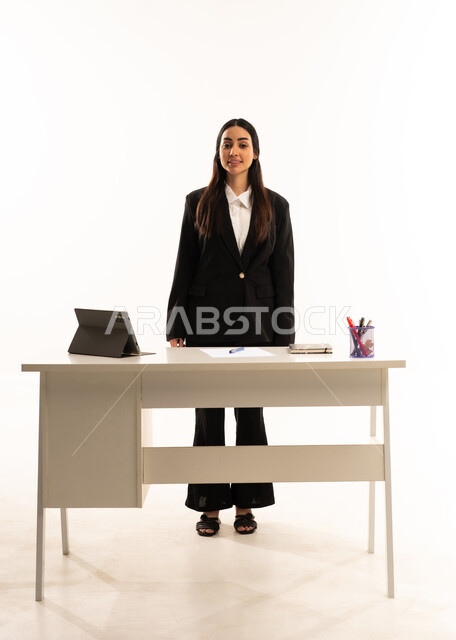 التميز في العمل, التعليم المتطور في مدارس المملكة العربية السعودية, بورتريه لمعلمه عربيه سعوديه خليجيه ترتدي البدله الرسميه السوداء تقف امام مكتب ابيض و تستخدم جهاز التابلت, ايماءات الثقة و السعادة, خلفية بيضاء