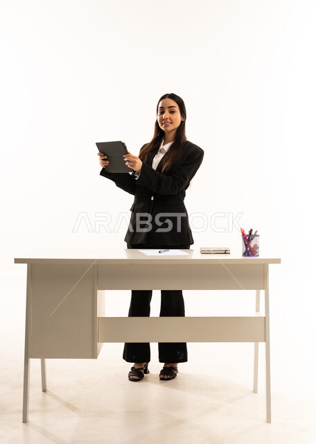 Promoting science, innovation, and skills development; advanced education in Saudi Arabian schools; portrait of a Saudi Arabian teacher wearing a black formal suit, standing in front of a white desk and holding a tablet; white background.