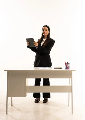 تعزيز العلوم والإبتكارات وتنمية المهارات، التعليم المتطور في مدارس المملكة العربية السعودية, بورتريه لمعلمه عربيه سعوديه خليجيه ترتدي البدله الرسميه السوداء تقف امام مكتب ابيض و تحمل جهاز التابلت, خلفية بيضاء