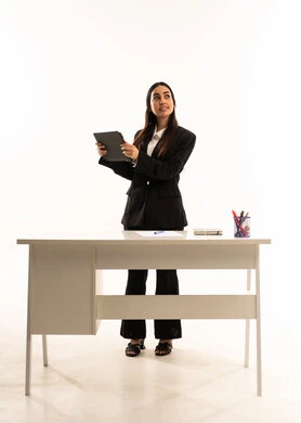 تنمية و تطوير المهارات، التعليم المتطور في مدارس المملكة العربية السعودية, بورتريه لمعلمه عربيه سعوديه خليجيه ترتدي البدله الرسميه السوداء تقف امام مكتب ابيض و تحمل جهاز التابلت, خلفية بيضاء