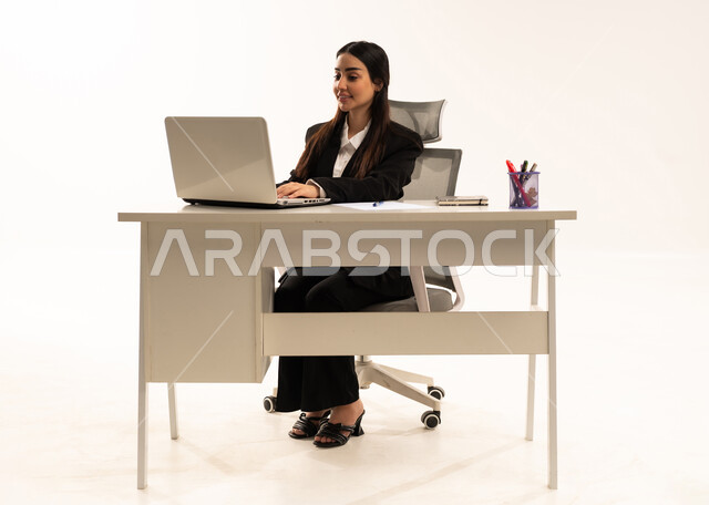 إنجاز المهام المطلوبة أونلاين، دمج العمل مع التكنولوجيا الحديثة، إجراء الاعمال المكتبيه داخل مقر الشركه عبر اللابتوب، بورتريه لامرأة عربية سعودية خليجية ترتدي البدلة الرسمية السوداء و تستخدم الحاسب المحمول, خلفية بيضاء