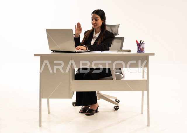 إيماءات يدين تدل على الترحيب, الاعتناء بالمظهر الخارجي، دمج العمل مع التكنولوجيا الحديثة، بورتريه لامرأة عربية سعودية خليجية ترتدي البدلة الرسمية السوداء و تتواصل مع العملاء من خلال اللابتوب, خلفية بيضاء