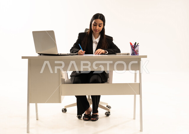 تدوين وتسجيل الملاحظات والمعلومات على الورق,  إجراء الاعمال المكتبيه داخل مقر الشركه، بورتريه لامرأة عربية سعودية خليجية ترتدي البدلة الرسمية السوداء تجلس خلف المكتب و أمامها جهاز اللابتوب، دراسه المشاريع ومراجعه التقارير السنويه، خلفية بيضاء