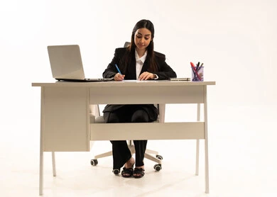 تدوين وتسجيل الملاحظات والمعلومات على الورق,  إجراء الاعمال المكتبيه داخل مقر الشركه، بورتريه لامرأة عربية سعودية خليجية ترتدي البدلة الرسمية السوداء تجلس خلف المكتب و أمامها جهاز اللابتوب، دراسه المشاريع ومراجعه التقارير السنويه، خلفية بيضاء