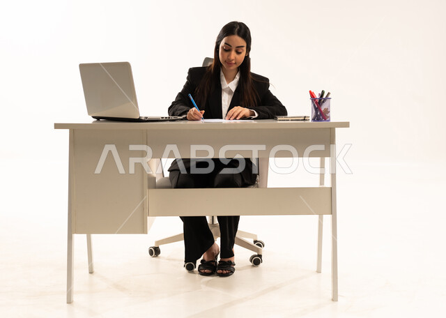 تدوين البيانات والملاحظات، إجراء الاعمال المكتبيه داخل مقر الشركه، بورتريه لامرأة عربية سعودية خليجية ترتدي البدلة الرسمية السوداء تجلس خلف المكتب و أمامها جهاز اللابتوب، دراسه المشاريع ومراجعه التقارير السنويه، خلفية بيضاء