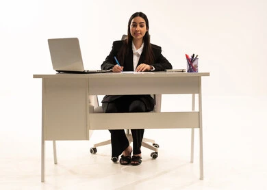 Writing job descriptions and objectives, performing office work within the company headquarters, a portrait of a Saudi Arabian woman from the Gulf wearing a black formal suit, sitting behind a desk with a laptop in front of her, studying projects and reviewing annual reports, white background