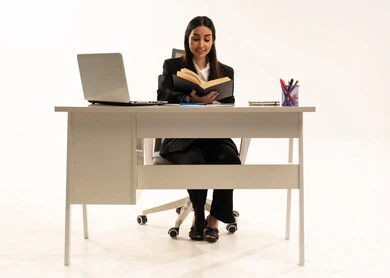 Reading and acquiring knowledge, studying and researching the project, performing office tasks and work within the company headquarters; a portrait of a Saudi Arabian Gulf woman wearing a black formal suit, sitting behind a desk and reading with focused and thoughtful gestures, white background.