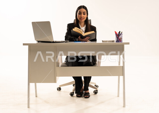 التدقيق في القراءة, المطالعة و دراسة المشروع، إجراء المهام والاعمال المكتبيه داخل مقر الشركه، بورتريه لامرأة عربية سعودية خليجية ترتدي البدلة الرسمية السوداء تجلس خلف المكتب و تقرأ بإيماءات التركيز والتمعن، خلفية بيضاء