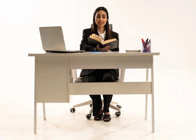 Reading, studying, and reviewing the project; performing office tasks and work within the company headquarters; a portrait of a Saudi Arabian Gulf woman wearing a black formal suit, sitting behind a desk and reading with focused and thoughtful gestures; white background.