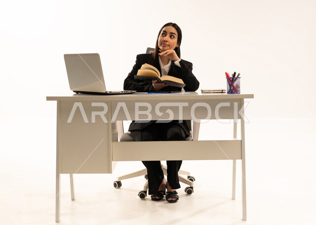 الاهتمام بتصفح الكتب المفيدة، القراءة والتزود بالعلم، إجراء المهام والاعمال المكتبيه داخل مقر الشركه، بورتريه لامرأة عربية سعودية خليجية ترتدي البدلة الرسمية السوداء تجلس خلف المكتب و تقرأ بإيماءات التركيز والتمعن، خلفية بيضاء