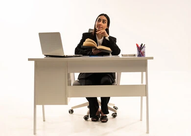 الاهتمام بتصفح الكتب المفيدة، القراءة والتزود بالعلم، إجراء المهام والاعمال المكتبيه داخل مقر الشركه، بورتريه لامرأة عربية سعودية خليجية ترتدي البدلة الرسمية السوداء تجلس خلف المكتب و تقرأ بإيماءات التركيز والتمعن، خلفية بيضاء