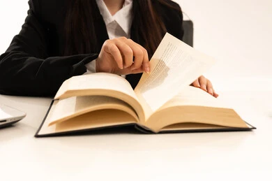 A close-up photograph of the hand of a Saudi Arabian woman from the Gulf region, wearing a black formal suit and browsing a book; reading and acquiring knowledge, studying and researching a project; white background.