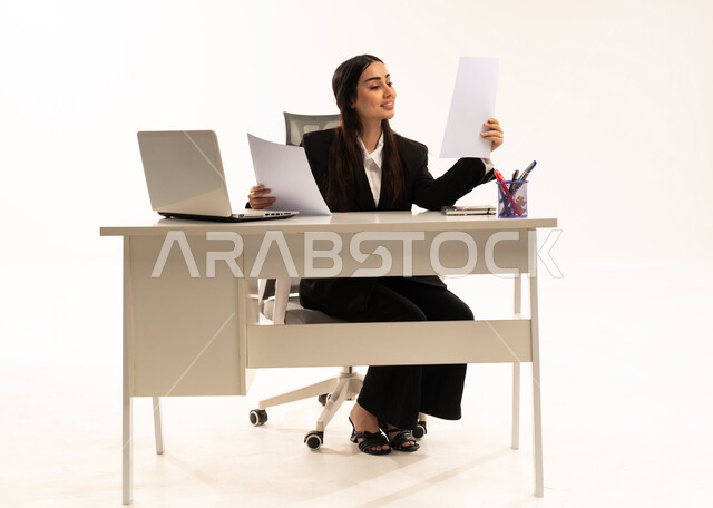 قراءة البنود الخاصة بالعقد، الانهماك في العمل, إجراء المهام والاعمال المكتبيه داخل مقر الشركه، بورتريه لامرأة عربية سعودية خليجية ترتدي البدلة الرسمية السوداء تجلس خلف المكتب و تحمل الأوراق, دراسه المشاريع و مراجعه التقارير السنويه، خلفية بيضاء
