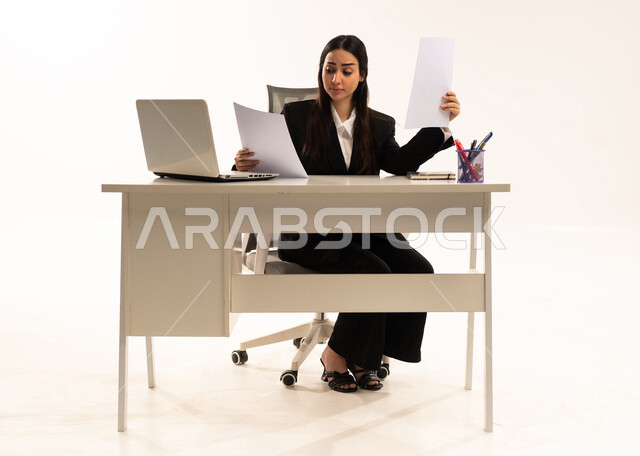الانهماك في العمل,  إجراء المهام والاعمال المكتبيه داخل مقر الشركه، بورتريه لامرأة عربية سعودية خليجية ترتدي البدلة الرسمية السوداء تجلس خلف المكتب و تحمل الأوراق, دراسه المشاريع و مراجعه التقارير السنويه، خلفية بيضاء