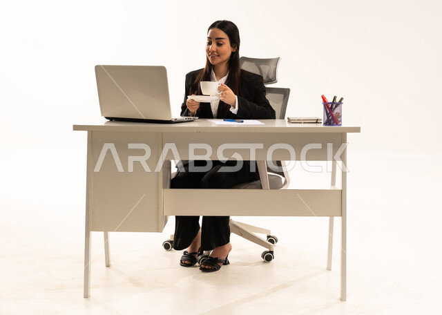 Drinking coffee while browsing social media, performing office tasks and work within the company headquarters; a portrait of a Saudi Arabian woman from the Gulf region wearing a black formal suit, sitting behind a desk working on a laptop, studying projects and reviewing annual reports, white background.