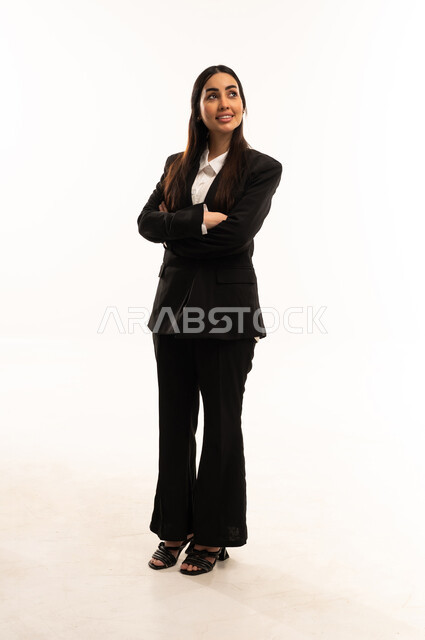 The concept of femininity and softness, attention to elegance and outward appearance, gestures of self-confidence, a portrait of a smiling Saudi Arabian Gulf woman wearing a black formal suit, standing upright and poised with her hands crossed, white background