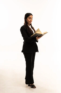 The concept of a passion for reading, the practice of reading as a hobby, gestures of focus and enjoyment, spending leisure time learning and self-education, a portrait of a smiling Saudi Arabian Gulf woman wearing a black formal suit, standing upright and holding a book, white background