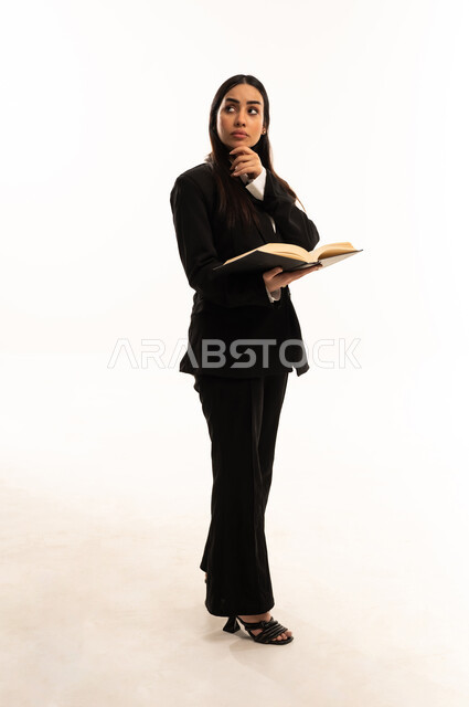 Reading enjoyable novels and stories, practicing the hobby of reading, gestures of focus and enjoyment, spending free time learning and self-education, a portrait of a smiling Saudi Arabian Gulf woman wearing a black formal suit, standing upright and holding a book, white background