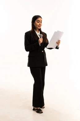 Conducting workshops and training courses, holding dialogues and discussions, delivering speeches and presentations using a microphone, starting voice training, portrait of a smiling Saudi Arabian Gulf woman wearing a black formal suit, standing upright and holding a microphone and papers, white background