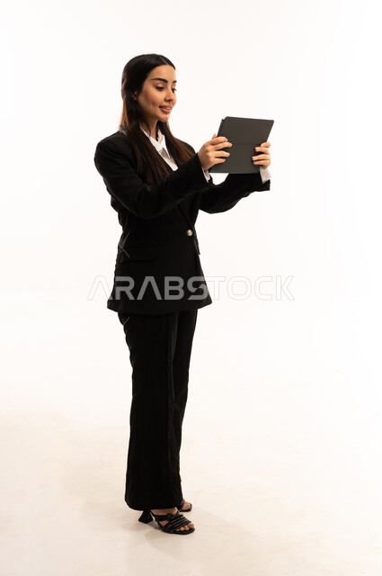 Utilizing modern techniques and advanced technology in the workplace, using a tablet to complete projects. Portrait of a smiling Saudi Arabian woman from the Gulf region, wearing a black formal suit, standing upright, holding a tablet and tapping on the screen. Attention to appearance, white background.