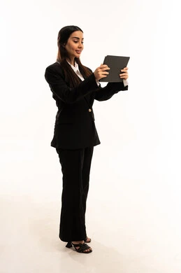 الاستفادة من التقنيات الحديثة والتكنولوجيا المتطورة في العمل, استخدم جهاز التابلت لانجاز المشاريع, بورتريه لامرأة عربية سعودية خليجية مبتسمة ترتدي البدلة الرسمية السوداء تقف باستقامة و تحمل الجهاز اللوحي و تقوم بالنقر على الشاشة, الاعتناء بالمظهر الخارجي، خلفية بيضاء