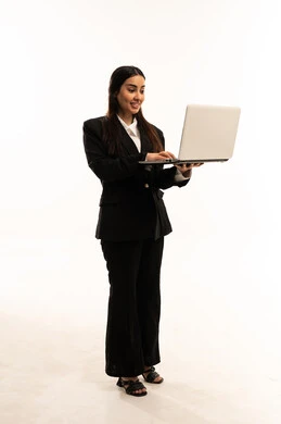 شرح خطوات العمل, استخدام اللابتوب في تنظيم الخطط، استخدام الأجهزة التقنية الحديثة في إنجاز المهام، بورتريه لامرأة عربية سعودية خليجية مبتسمة ترتدي البدلة الرسمية السوداء تقف باستقامة و تستخدم الحاسب المحمول, خلفية بيضاء
